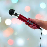 A person's hand holds a Doxy Die Cast 3 Wand Massager in Candy Red, showcasing its black rounded tip and silver accents, with a blurred background of colorful bokeh lights.